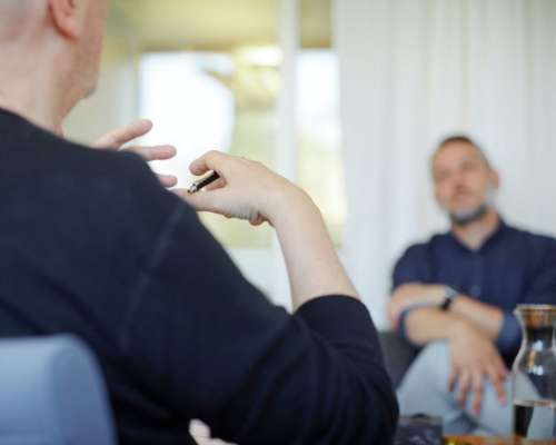 A man sits with his legs crossed while listening to a person speaking across the room