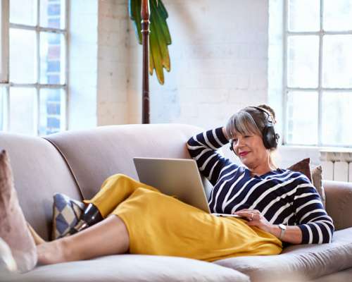 An older woman lays across a couch with headphones on and watches her computer