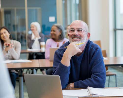 A man in a classroom smiles while watching his teacher